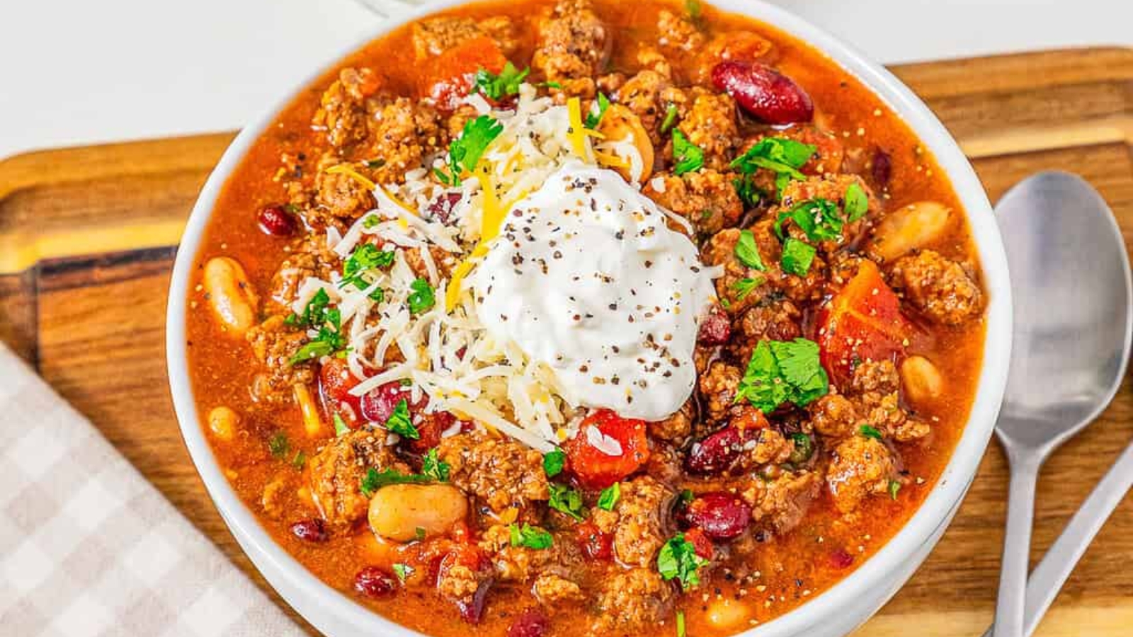 A bowl of chili topped with shredded cheese, sour cream, chopped parsley, and cracked black pepper.