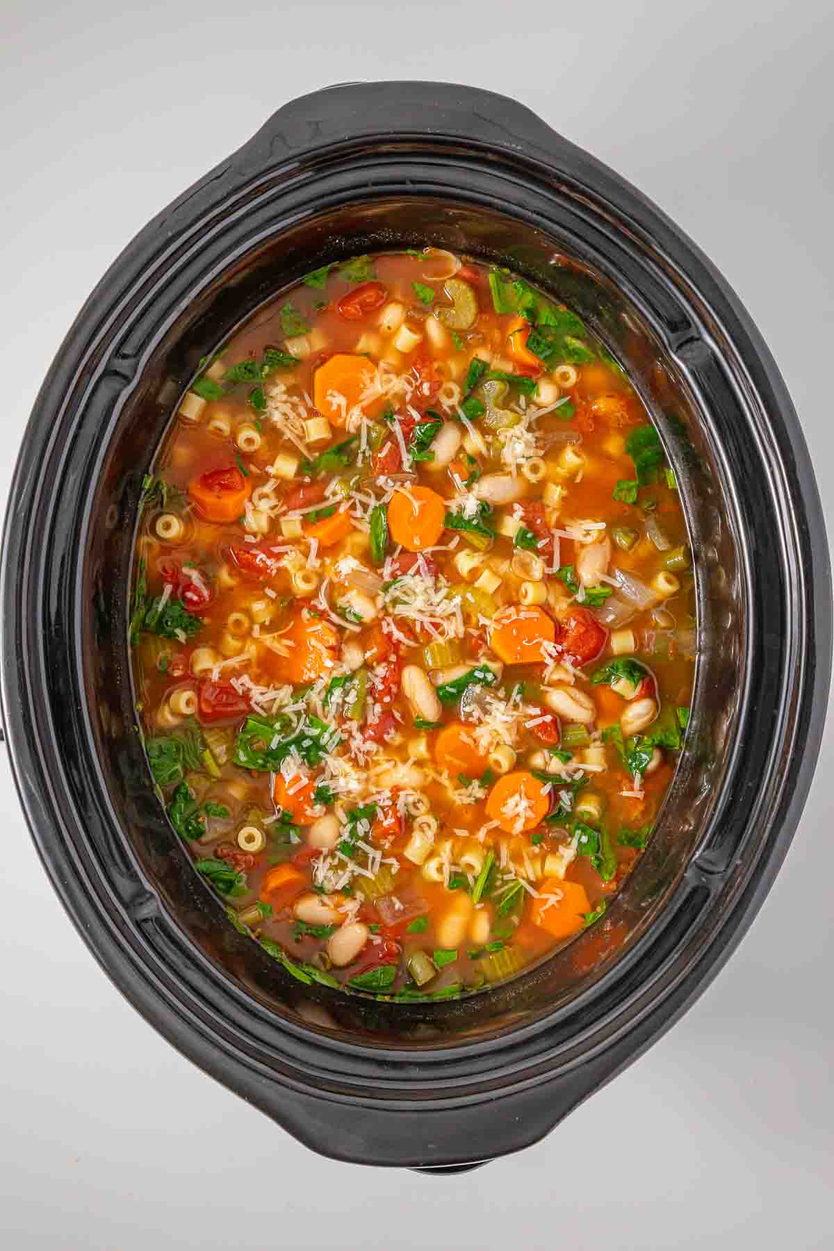 A slow cooker filled with vegetable and pasta minestrone soup topped with shredded cheese.