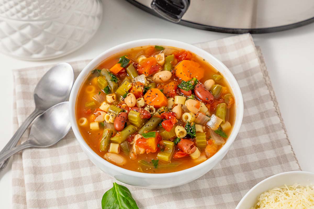 A bowl of slow cooker vegetable minestrone soup with pasta and beans on a checkered napkin.
