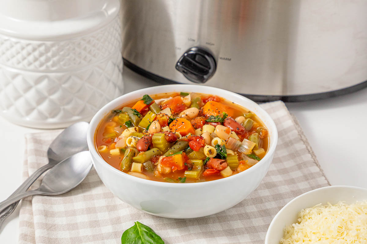 A bowl of Slow Cooker Minestrone soup next to a slow cooker and a side of grated cheese.