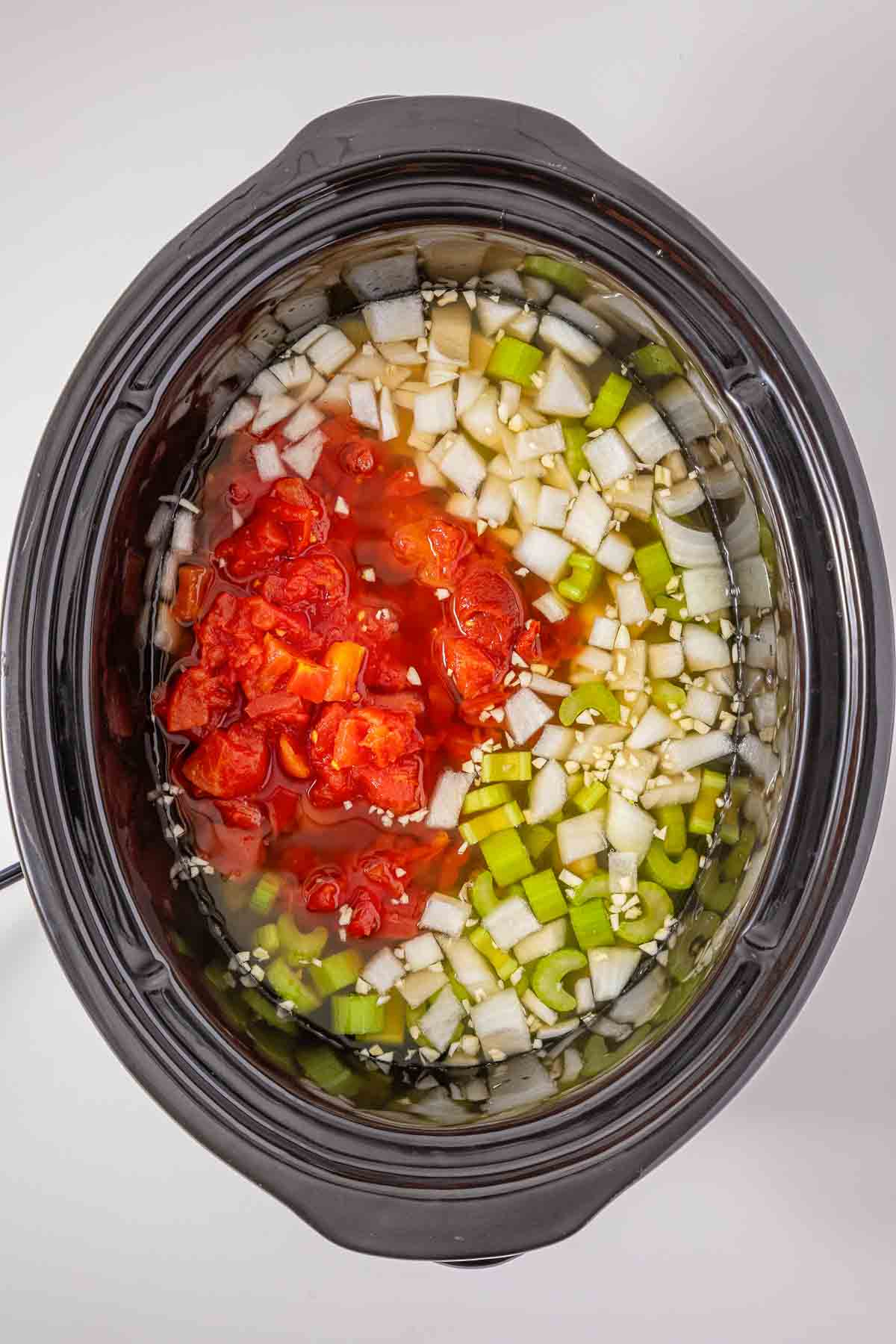 Slow cooker filled with ingredients for minestrone soup, including diced tomatoes, onions, celery, and garlic in liquid.
