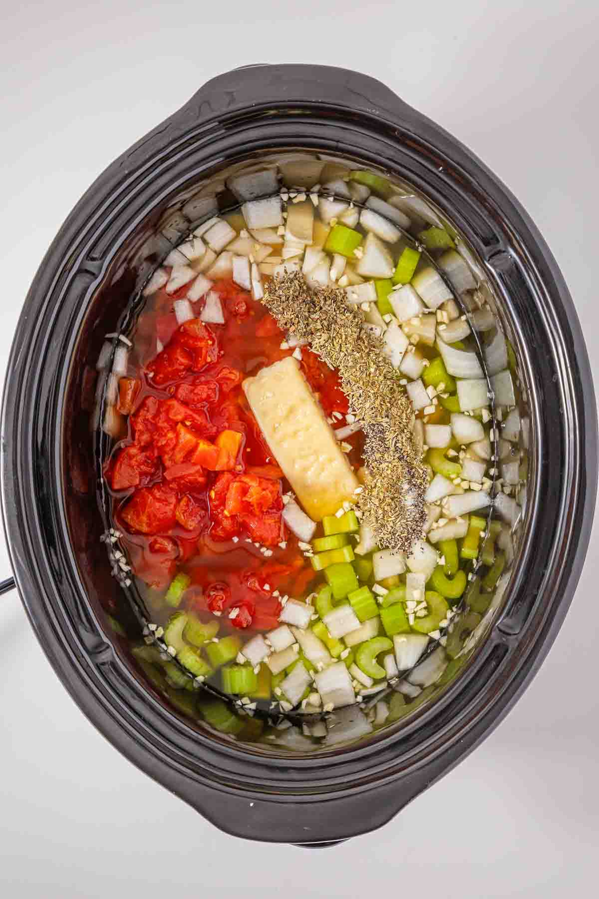 Slow cooker filled with ingredients for a minestrone soup, including onions, celery, tomatoes, butter, and dried herbs.