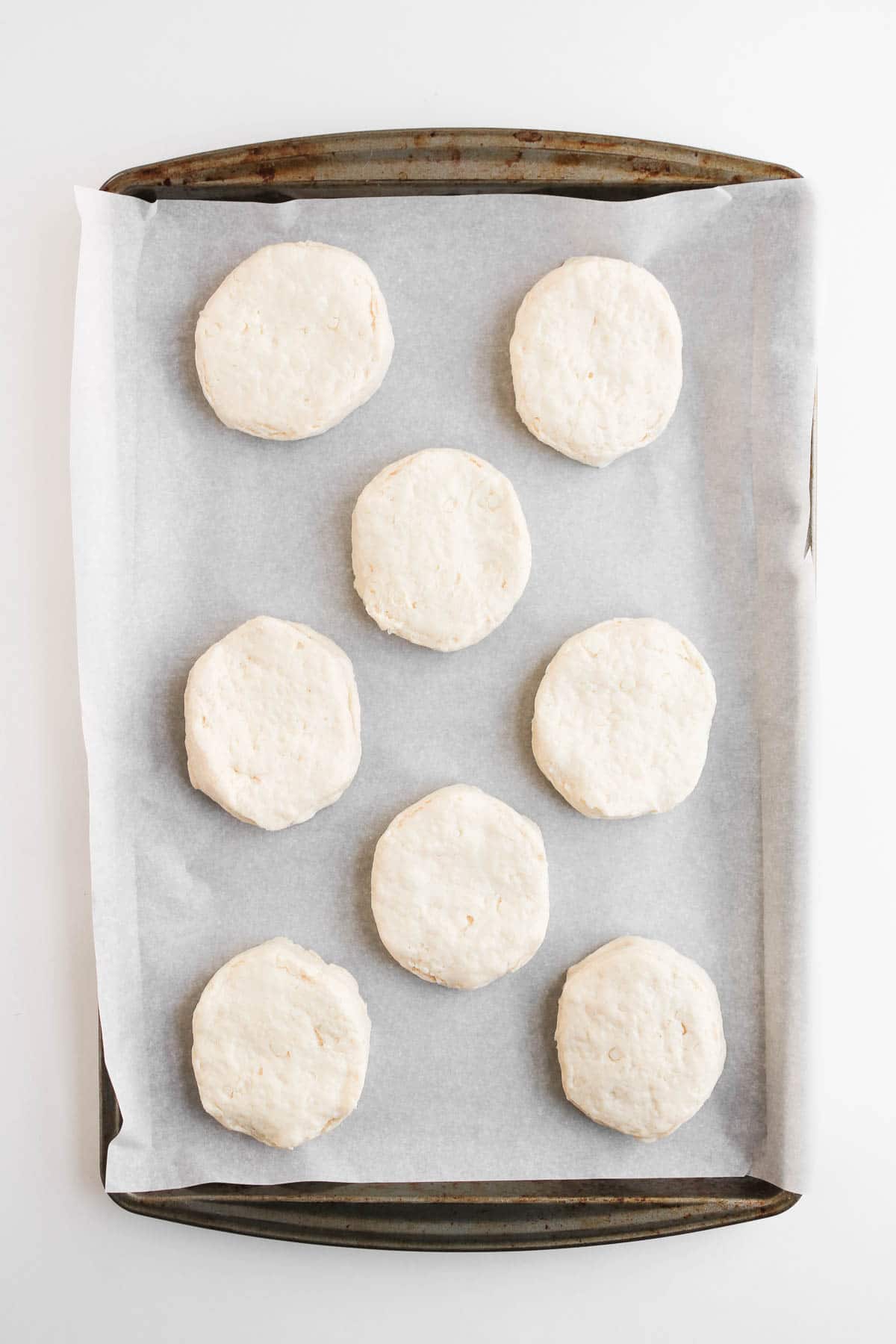Seven raw biscuit dough rounds for chicken pot pie arranged on a parchment-lined baking sheet.