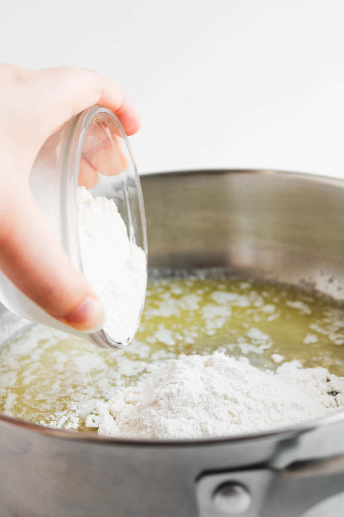 Hand pouring flour into melted butter to thicken the base for a chicken pot pie with biscuits.