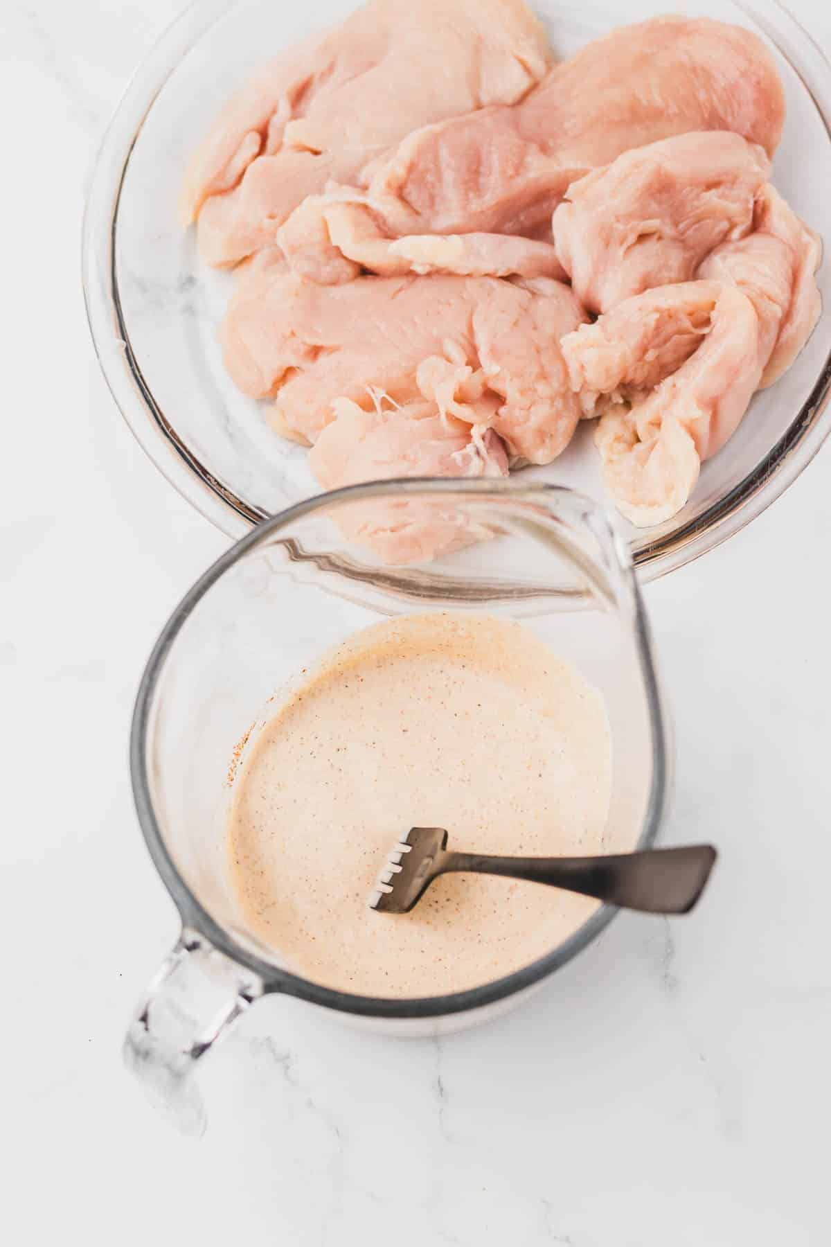 Raw chicken breasts in a glass bowl next to a whisk and creamy sauce for Copycat Longhorn Parmesan Crusted Chicken.