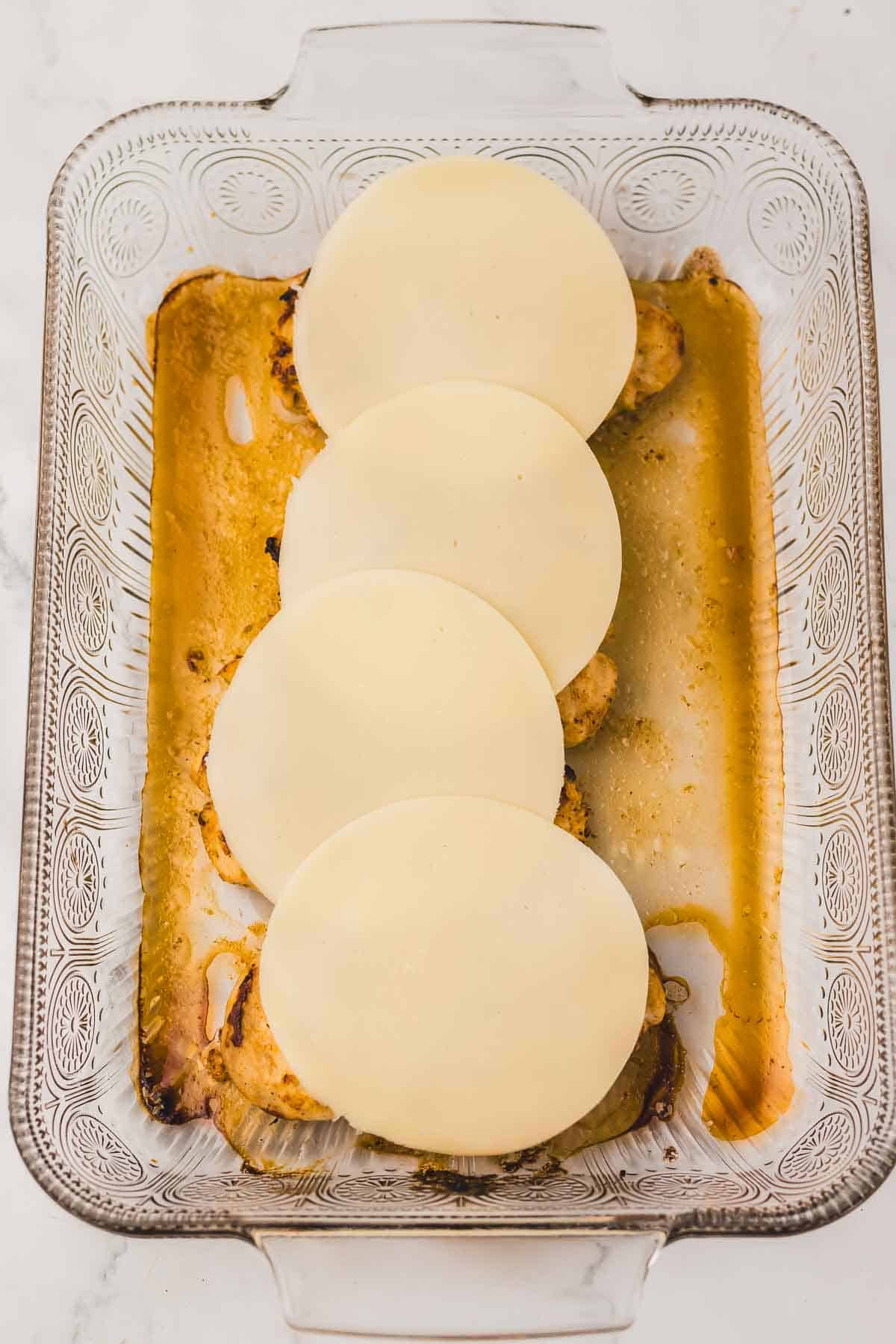Four slices of cheese layered over Copycat Longhorn Parmesan Crusted Chicken in a glass baking dish.