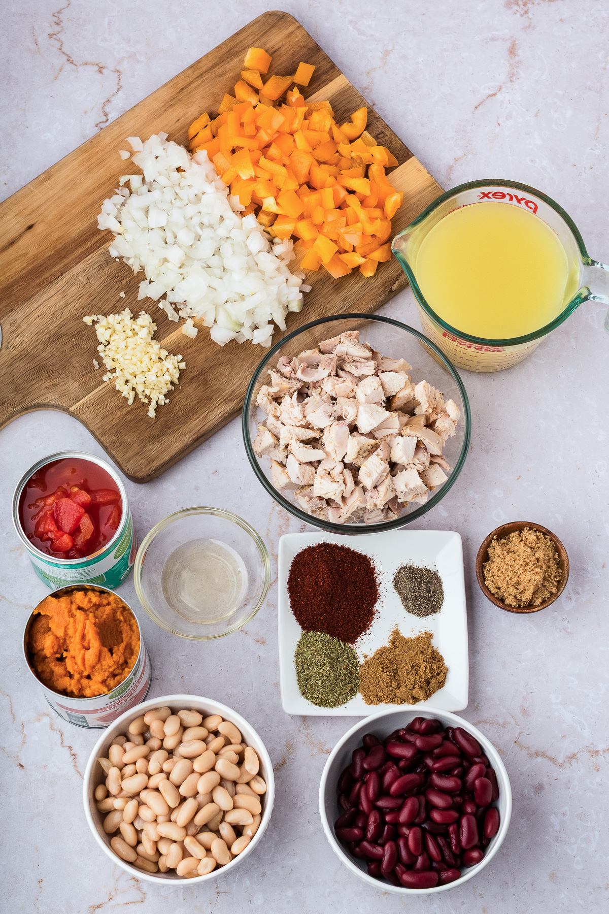 Ingredients for chili including chopped vegetables, diced chicken, beans, spices, tomato, pumpkin, and broth.