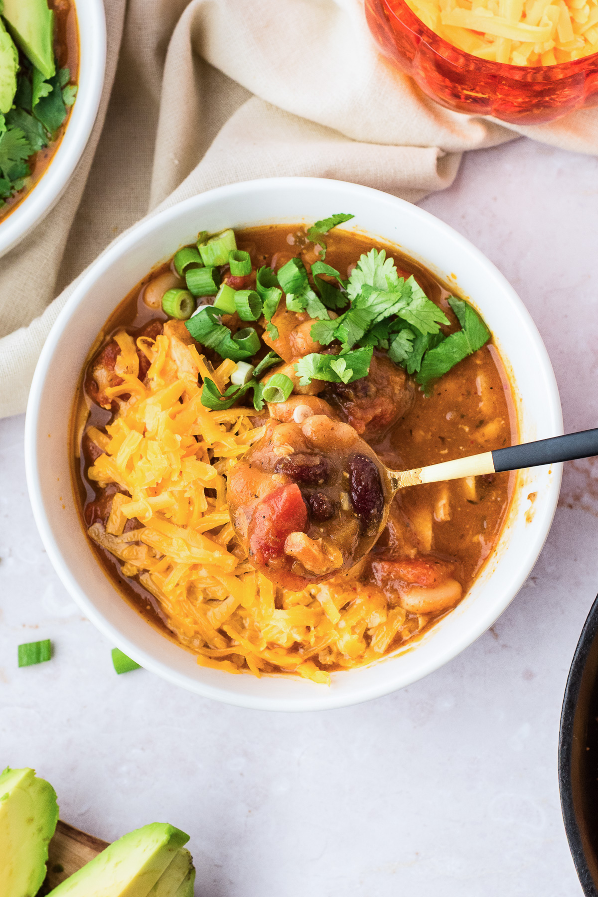 A bowl of leftover turkey pumpkin chili topped with shredded cheese, green onions, and cilantro, with a spoonful lifted.
