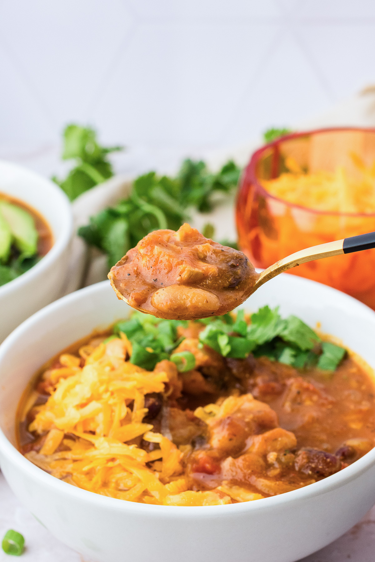 A spoonful of leftover turkey pumpkin chili held over a bowl topped with shredded cheddar cheese and cilantro.