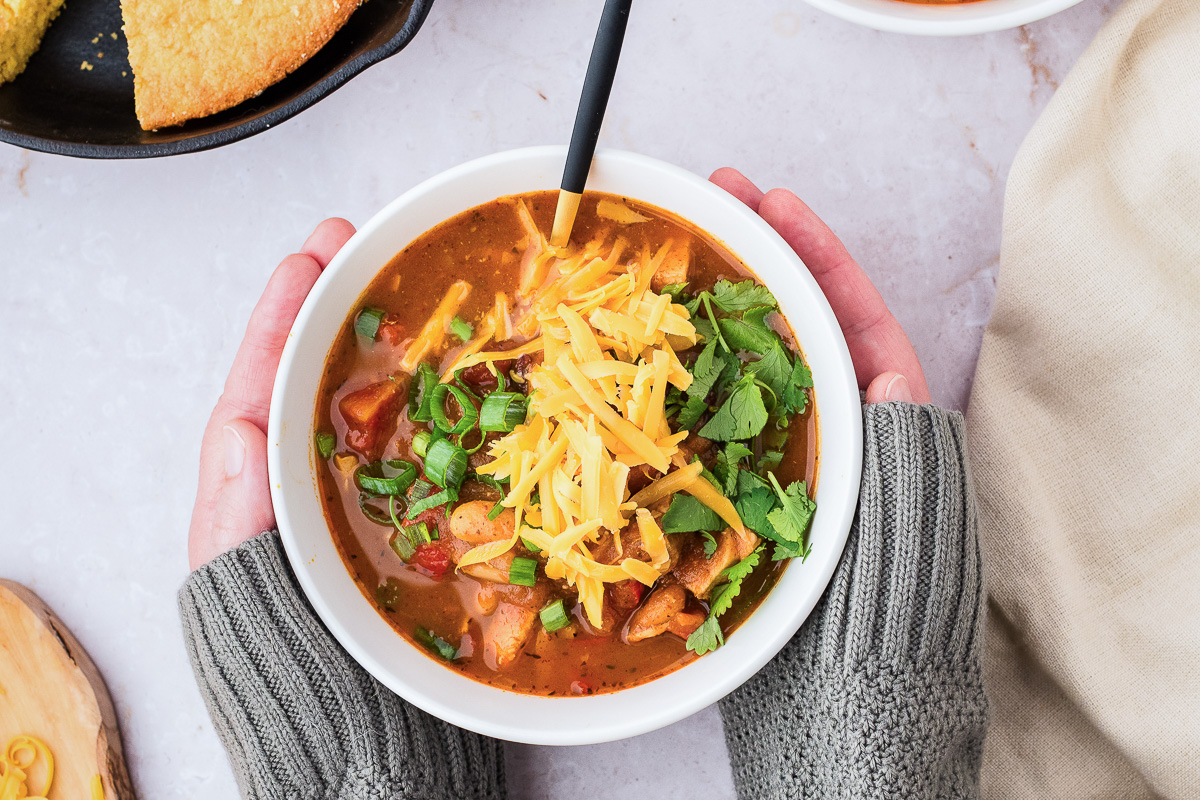Hands in gray sweaters holding a bowl of leftover turkey pumpkin chili topped with cheese, green onions, and cilantro.
