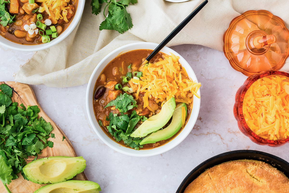 A bowl of leftover turkey pumpkin chili topped with shredded cheese, cilantro, and avocado slices.