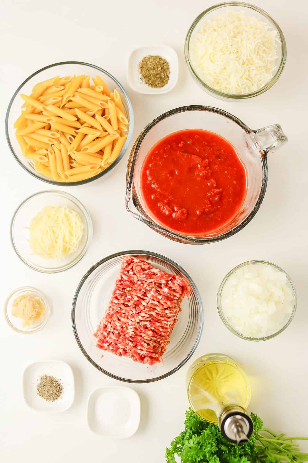 Penne, ground meat, and tomato sauce in glass bowls, prepped for a sausage pasta bake on a white surface.