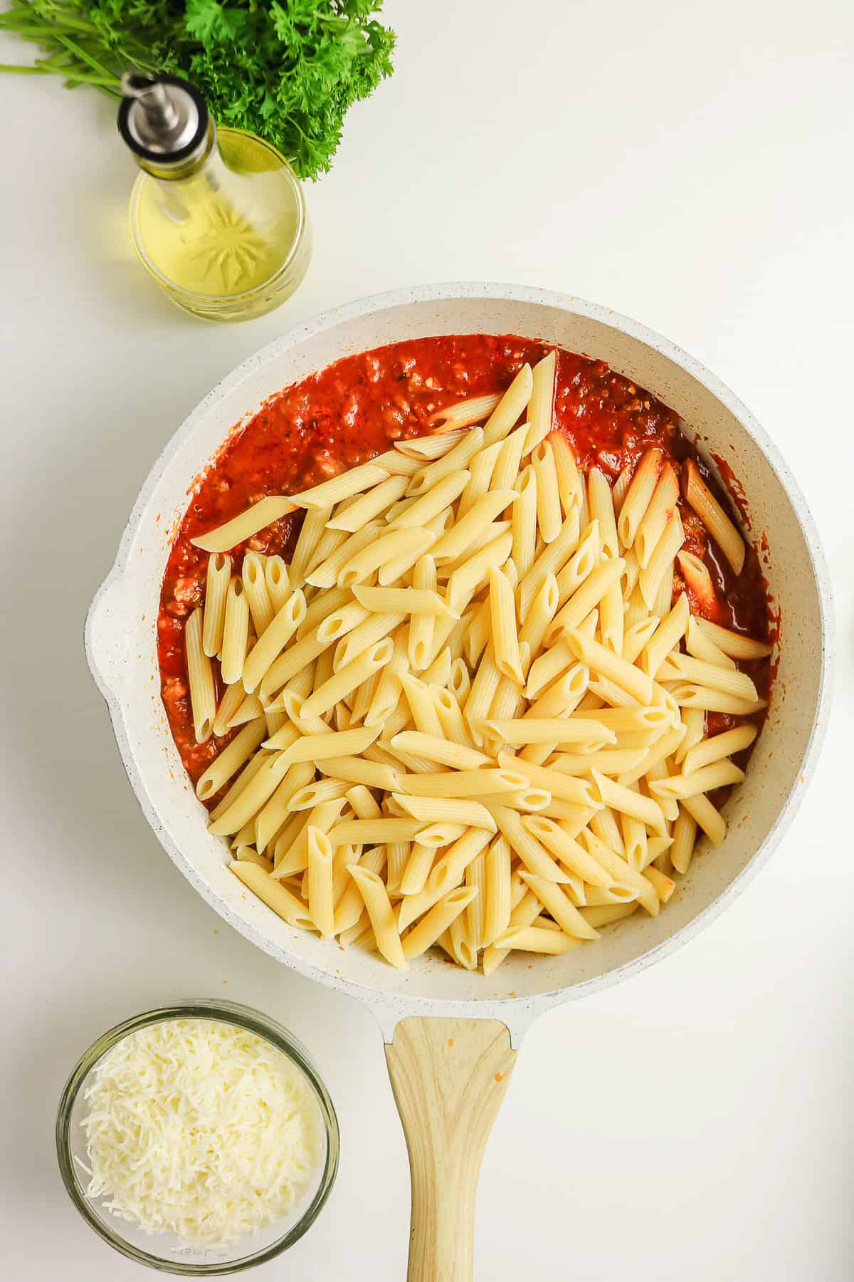 Penne pasta in a sausage pasta bake, with a bowl of shredded cheese and a bottle of oil nearby.