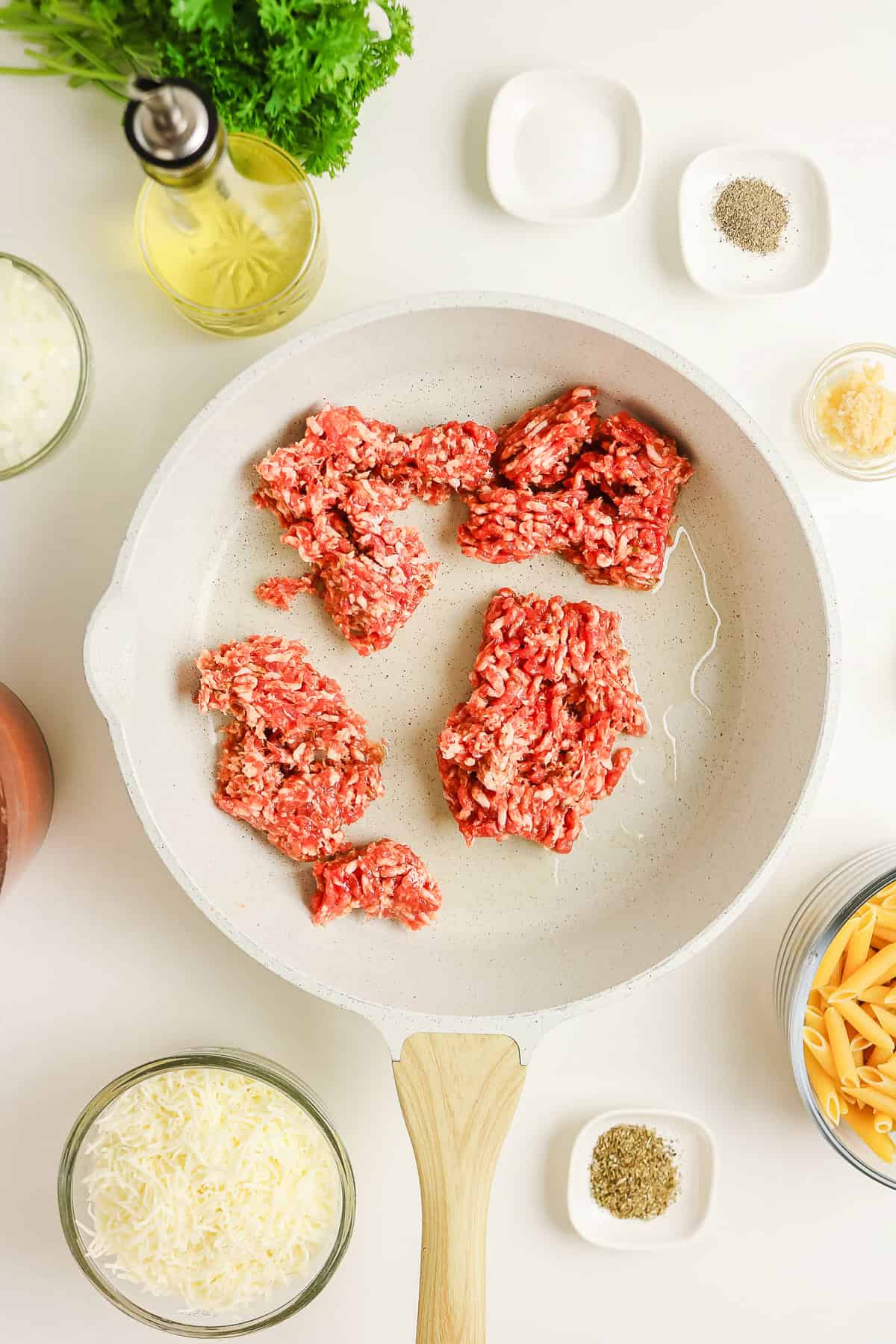 Sausage pasta meat cooking in a skillet surrounded by bowls of parsley, oil, garlic, and pasta.