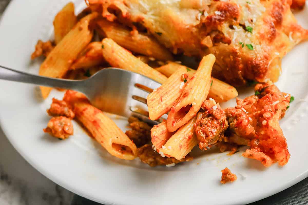 A fork lifting penne with meat sauce and a portion of sausage pasta bake on a white plate.