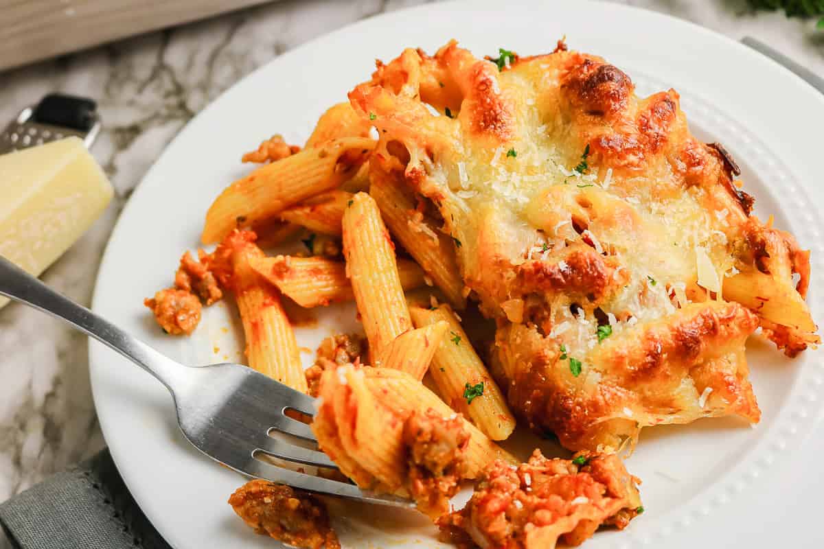 Baked sausage pasta with meat sauce and melted cheese on a white plate with a fork.