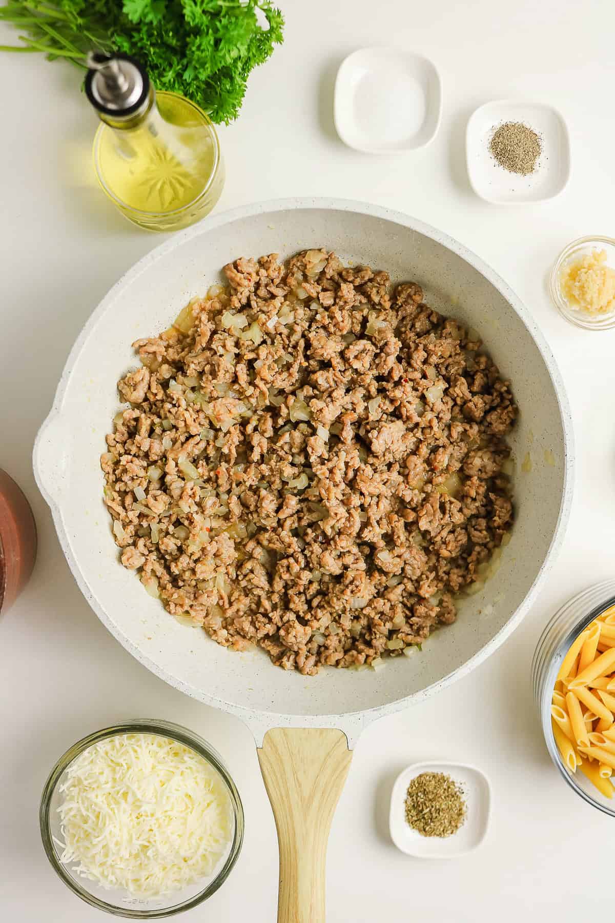 Ground meat and onions cook in a skillet for a sausage pasta bake, surrounded by bowls of pasta, cheese, and spices.