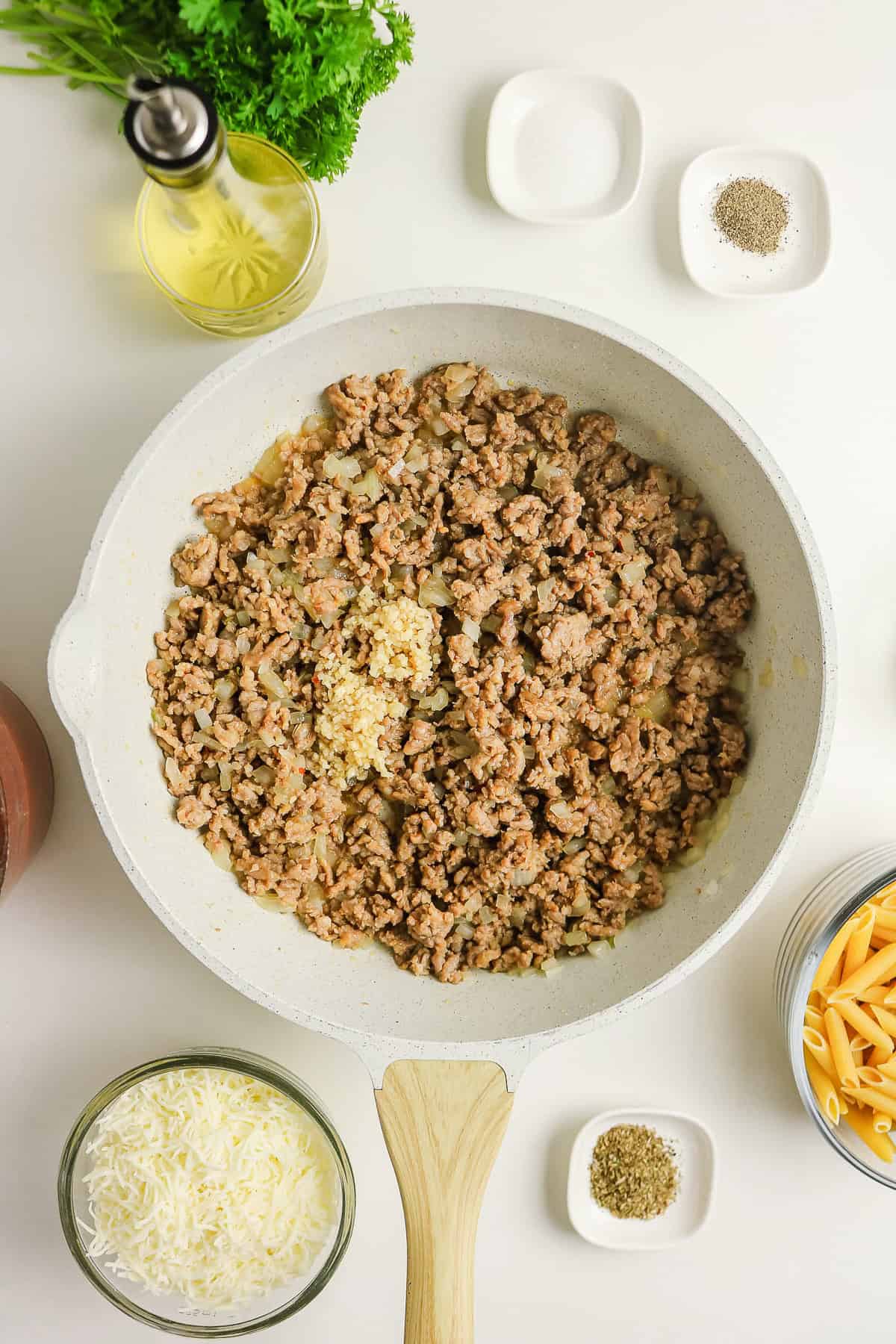Ground meat and onions cooking in a skillet to prepare a sausage pasta bake, with pasta, cheese, and spices nearby.
