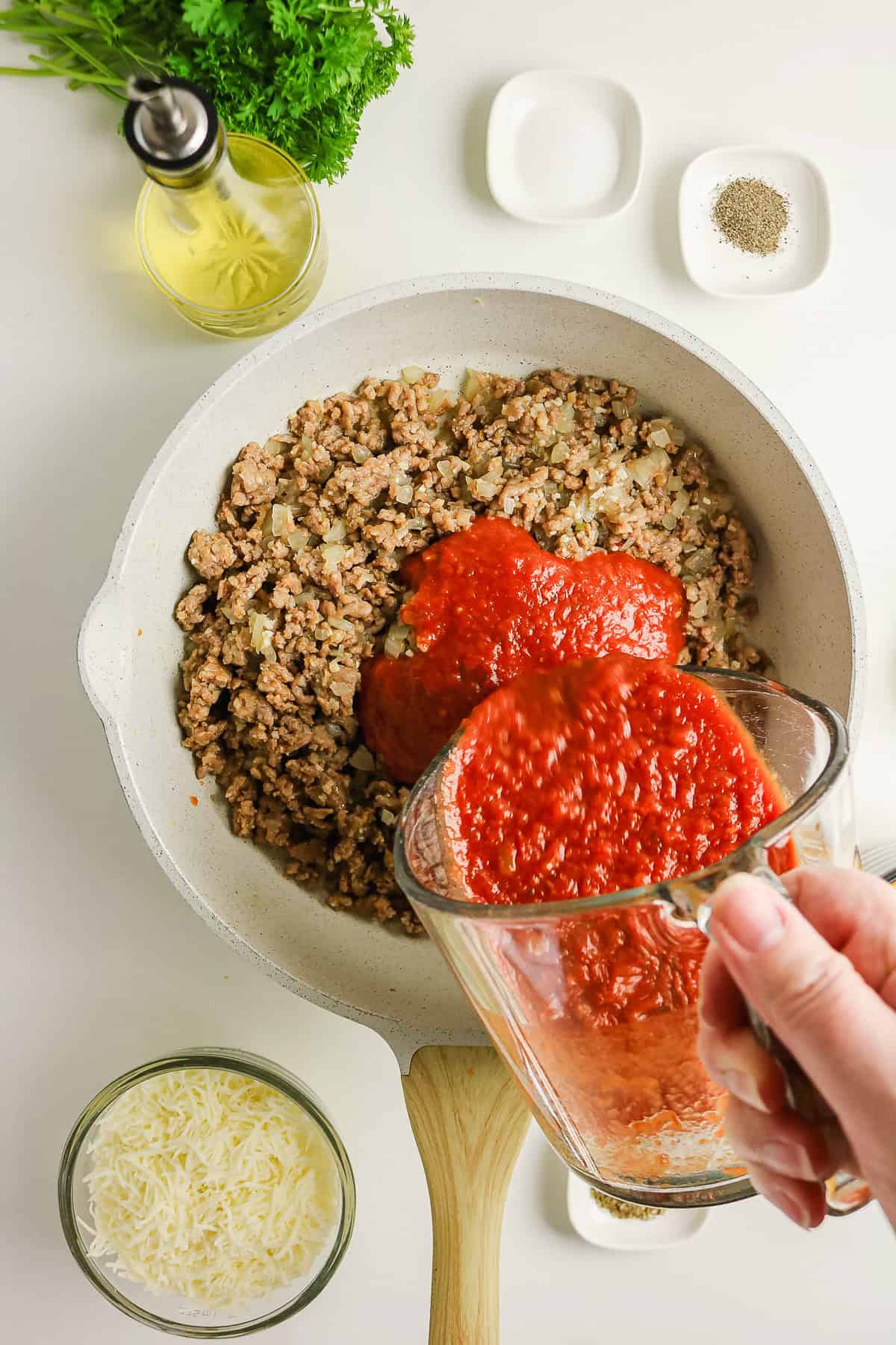 Tomato sauce poured from a glass measuring cup into a skillet of browned meat and onions for an easy pasta bake.