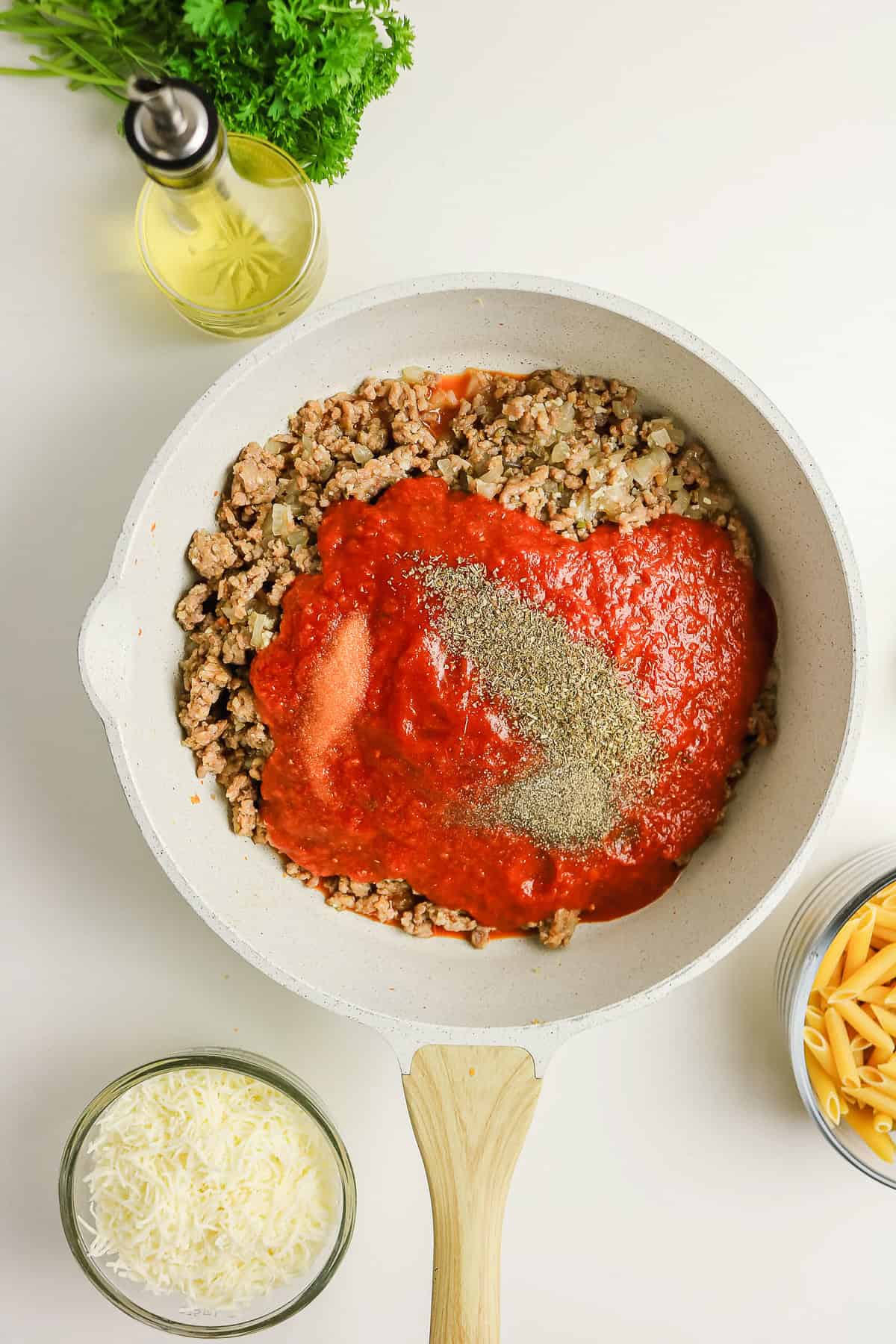 Ground meat, tomato sauce, and spices in a pan for a pasta bake recipe, surrounded by bowls of cheese, herbs, and oil.